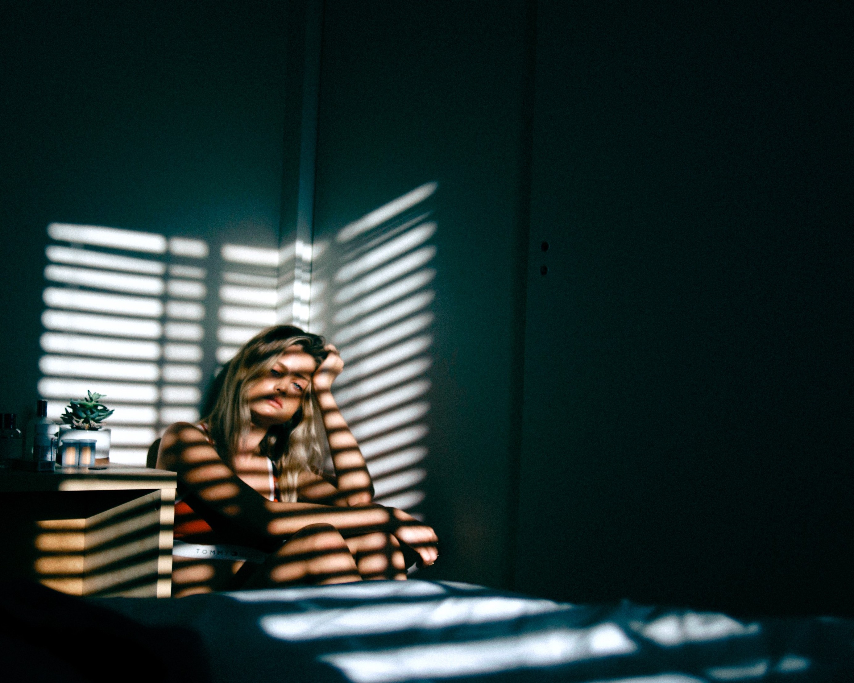 A dejected looking woman sat in a dark bedroom with her hand on her head, with horizontal strips of light shining on her from a blind, Bloomington.