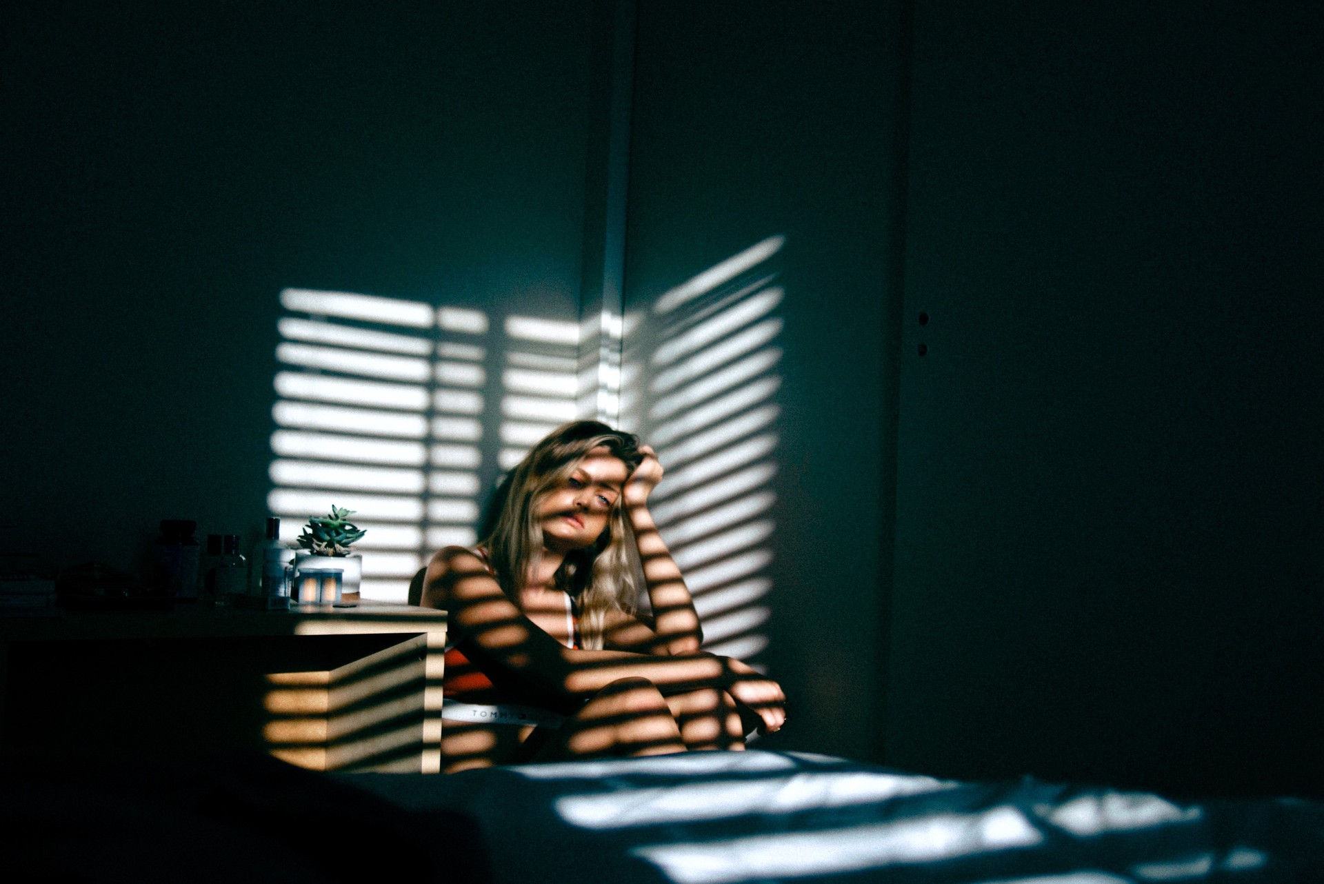 A dejected looking woman sat in a dark bedroom with her hand on her head, with horizontal strips of light shining on her from a blind, Bloomington.